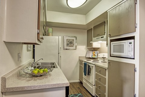 A compact kitchen here at Paradise Falls featuring light cabinetry, generous countertop space, and a functional layout with a sink, range, built-in microwave, and refrigerator, complemented by wood-style flooring and neutral finishes that create an efficient, well-organized space ideal for everyday cooking and meal preparation.