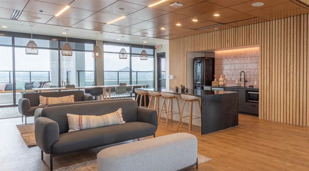 Modern open-concept lounge here at Joy on 4th Apartments with sleek sofas, a wooden ceiling, and island counter with barstools. Large windows offer a view of distant mountains.