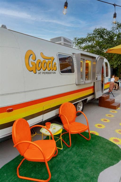 Vintage trailer with "Goods at Pemberton" branding near Joy on 4th Apartments, adorned with orange and yellow stripes. Two bright orange chairs sit outside on artificial grass.