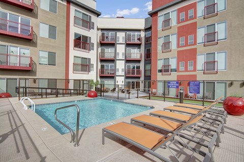 A modern outdoor pool courtyard here at Proxy 333 featuring a rectangular swimming pool with stainless handrails, surrounded by orange lounge chairs on a spacious concrete deck, set against contemporary apartment buildings with red-accented balconies, secure fencing, and shared seating areas creating a vibrant urban retreat.