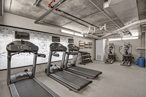 A modern fitness center here at Proxy 333 featuring multiple treadmills with mounted screens, elliptical machines, and a free weight rack set on durable flooring, with an industrial-style exposed concrete ceiling, bright overhead lighting, and clean neutral walls creating a functional and energizing workout space.