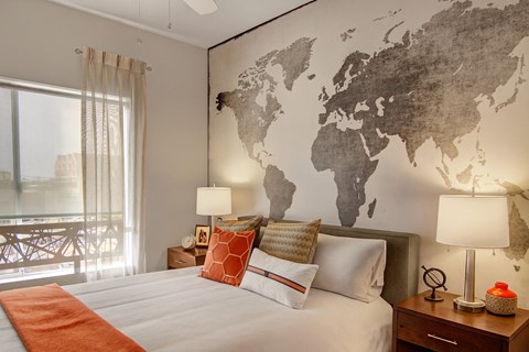 A stylish bedroom here at Proxy 333 featuring a comfortable bed dressed in crisp neutral bedding with layered accent pillows, matching nightstands with modern table lamps, a striking world map accent wall, soft window coverings, and a large window that brings in natural light for a warm and inviting retreat.