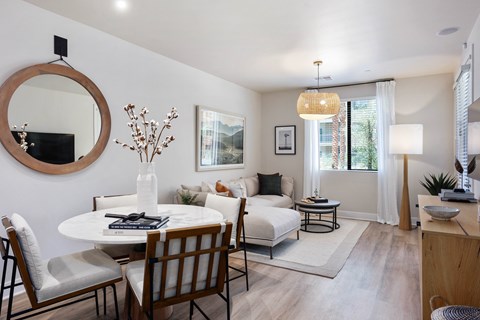 A bright and inviting open living and dining area here at Reflect at Dobson Ranch featuring a round dining table with upholstered chairs, a cozy sectional sofa with neutral and warm accent pillows, light wood-style flooring, layered rugs, soft window treatments, modern wall art, and natural light that creates a comfortable and welcoming atmosphere.