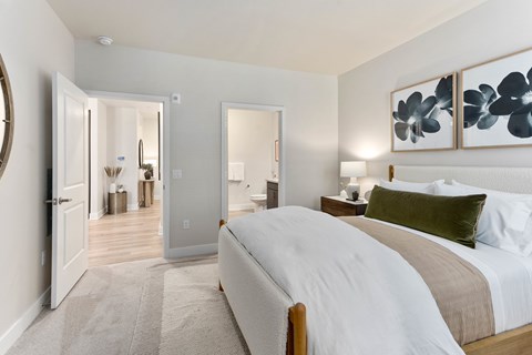 A tranquil bedroom here at Reflect at Dobson Ranch showcasing a plush upholstered bed with crisp white bedding, a soft neutral throw, and layered accent pillows, paired with warm wood nightstands and modern table lamps, with framed botanical artwork above the headboard, plush carpeting underfoot, and open doorways leading to an en suite bathroom and hallway.
