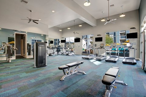A spacious fitness center here at Rio Paradiso featuring a wide open layout with treadmills, stationary bikes, weight machines, and adjustable benches arranged on patterned flooring, complemented by high ceilings with ceiling fans, wall-mounted televisions, and large windows that bring in natural light and views of the outdoor pool area.