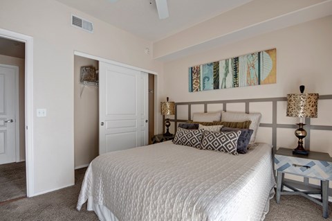 A serene bedroom here at Rio Paradiso featuring a spacious bed dressed in neutral bedding with patterned accent pillows, matching bedside tables with decorative lamps, and soft carpet flooring. The room includes a ceiling fan, an open closet with shelving, and a private doorway leading to an adjoining bathroom for added comfort and convenience.