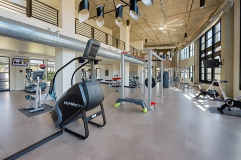 A spacious fitness center here at Roosevelt Row showcasing an open-concept gym with high ceilings, exposed ductwork, large windows for natural light, and a variety of cardio machines, strength equipment, and free weights arranged across polished flooring, creating a modern and motivating workout environment.