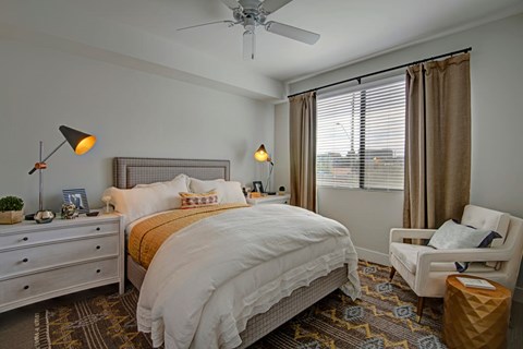 A serene bedroom here at Roosevelt Row featuring a plush upholstered bed dressed in layered neutral bedding, matching nightstands with modern accent lamps, a cozy armchair with a small side table, patterned area rug over wood-style flooring, a ceiling fan, and a large window with blinds and curtains allowing soft natural light to fill the space.