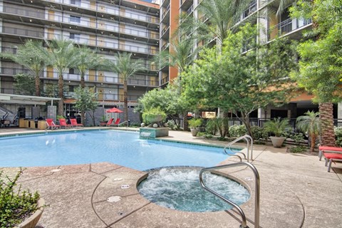 A resort-style outdoor pool here at Skyline Lofts featuring a sparkling swimming pool with an attached circular spa, curved concrete decking, and stainless steel handrails, surrounded by tall palm trees, lush desert landscaping, and red lounge chairs, all set within a modern courtyard framed by multi-level residential balconies.