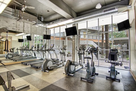 A modern fitness center here at Skyline Lofts featuring rows of cardio machines including treadmills, ellipticals, and stationary bikes, set on durable flooring beneath exposed concrete ceilings and ductwork, with wall-mounted TVs, ceiling fans, and floor-to-ceiling windows overlooking a landscaped courtyard with palm trees.