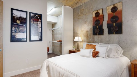 A modern loft-style bedroom here at Skyline Lofts featuring a comfortable bed with layered neutral bedding and accent pillows, exposed concrete walls with bold abstract wall art, a bedside table with a table lamp, soft carpeted flooring, and an open closet area with shelving that adds storage while maintaining the industrial-inspired aesthetic.