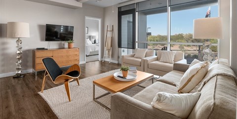 A bright living room here at Skywater at Tempe Town Lake featuring a neutral sofa with textured throw pillows, two coordinating armchairs, and a warm wood coffee table set on a plush area rug over wood-style flooring, accented by a sculptural chair, modern floor lamp, ceiling fan, and large windows that bring in abundant natural light and views of the surrounding landscape.