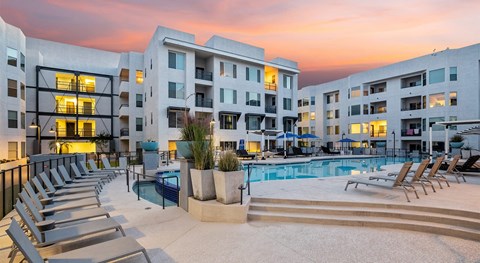 A resort-style courtyard pool here at Skywater at Tempe Town Lake showcasing a large swimming pool with a raised spa, tiered steps, and surrounding lounge chairs, framed by modern mid-rise apartment buildings, landscaped planters, and ambient lighting under a colorful sunset sky that creates a relaxing community atmosphere.