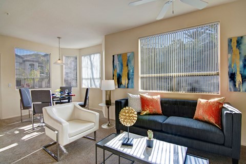 A welcoming living room here at Sonoma Landing featuring a dark upholstered sofa accented with warm orange pillows, a sleek glass coffee table with modern décor, and a white accent chair, while large windows with vertical blinds fill the space with natural light and connect to a dining area with a contemporary table, chairs, and pendant lighting.