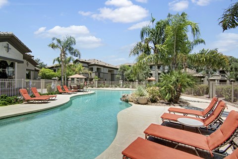 A resort-style outdoor swimming pool here at Sonoma Landing featuring a curved turquoise pool surrounded by red cushioned lounge chairs, palm trees, and desert landscaping, with shaded umbrellas, a fenced deck, and warm stucco apartment buildings creating a relaxing courtyard setting under a bright blue sky.