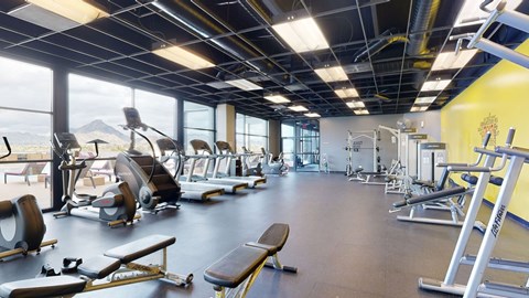 A spacious fitness center here at Trend at 51 featuring rows of treadmills and elliptical machines along floor-to-ceiling windows with mountain views, strength training equipment and benches arranged throughout, open rubber flooring, exposed ceiling details, and bright accent walls creating an energizing workout environment.