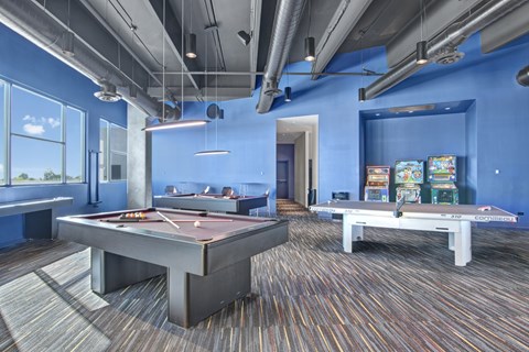 A vibrant resident game room here at Trend at 51 featuring multiple pool tables with overhead pendant lighting, a ping pong table, classic arcade machines along a bold blue accent wall, exposed industrial-style ceilings with visible ductwork, patterned carpeting, and large windows that bring in natural light and views of the surrounding area.