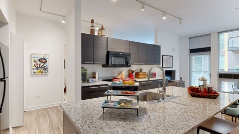 A modern open kitchen here at Trend at 51 showcasing a large granite island with an integrated sink and gooseneck faucet, dark flat-panel cabinetry, and sleek built-in appliances, complemented by track lighting, light wood-style flooring, and an open sightline into the adjacent living area through tall windows with blinds.