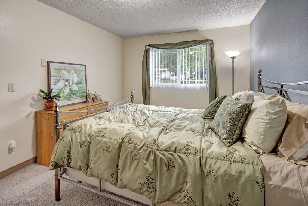 Cozy bedroom here at Brighton Wood Apartments with a green bedspread and matching pillows, a wooden dresser with decorative items, and a window with green curtains, creating a serene vibe.