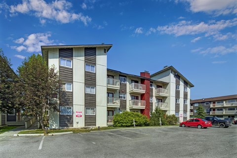 A red car is parked in a parking lot in front of a multi-story apartment building.