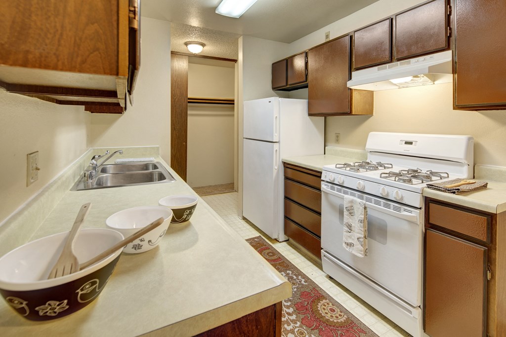 Narrow kitchen here at Taiga Apartments with brown cabinets, white appliances including a refrigerator and gas stove, three bowls on the counter, and a patterned rug on the floor.
