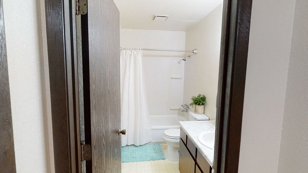 View from an open doorway into a small bathroom here at Deer Park Apartments featuring a white shower curtain, a sink with a marble top, and a potted plant.