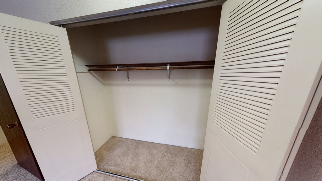 Interior view of an empty closet here at Deer Park Apartments with a carpeted floor, a brown shelf, and a hanging rod for clothes, flanked by paneled doors.