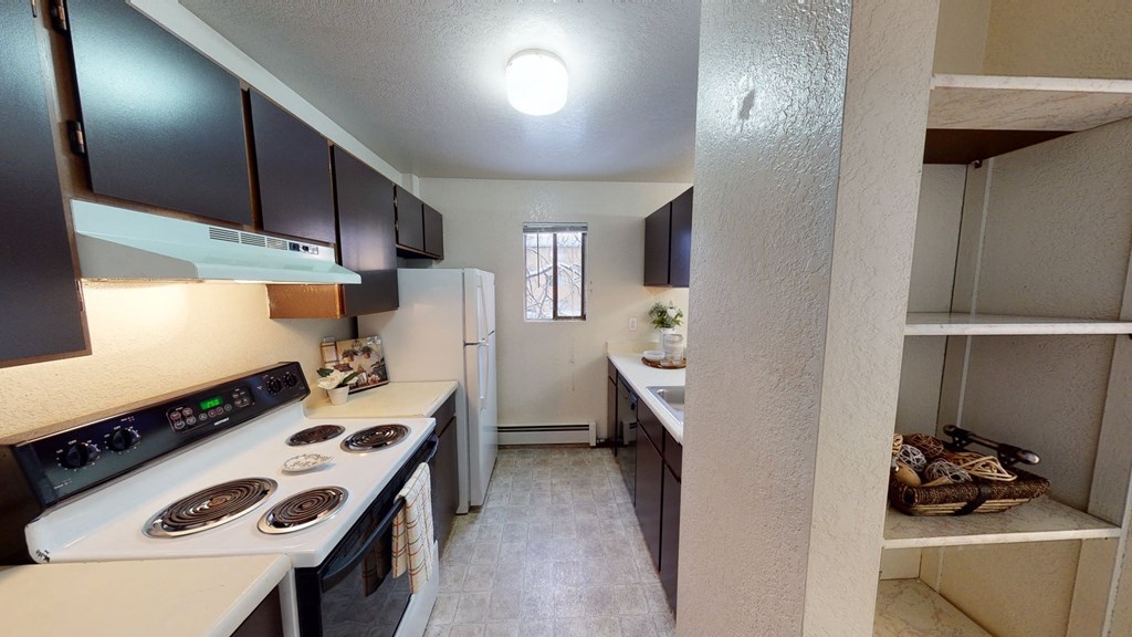 Narrow kitchen here at Deer Park Apartments with dark cabinets, white appliances, and tiled floor. Bright overhead light, window, and decorative shelf enhance a cozy feel.