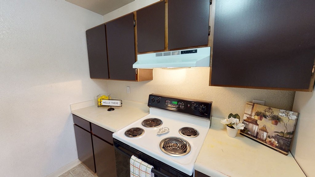 Cozy kitchen here at Deer Park Apartments with dark brown cabinets, white stove, and beige countertops. Cheerful decor includes a "Hello There" sign and a small plant.