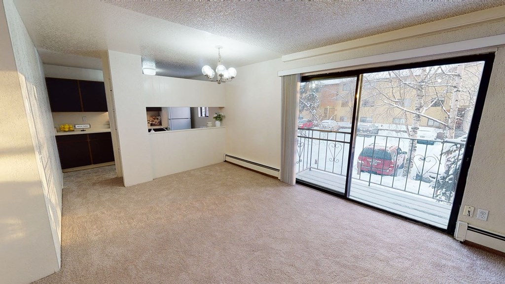Cozy apartment interior here at Deer Park Apartments with beige carpet, a small kitchen with dark cabinets, and large sliding glass doors leading to a snowy balcony view.