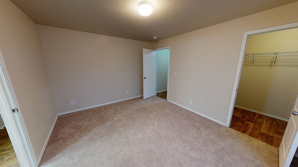 Beige Bedroom here at Four Seasons Apartments with carpeted floor, lit by a ceiling light. Open doors reveal a hallway with wooden flooring and a closet with wire shelving.