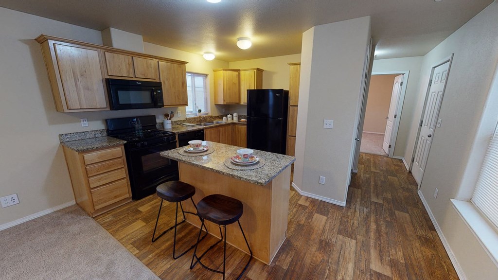 Bright kitchen here at Four Seasons Apartments with wooden cabinets, granite countertops, and a black appliance set. Two wooden stools at an island create a cozy, welcoming ambiance.