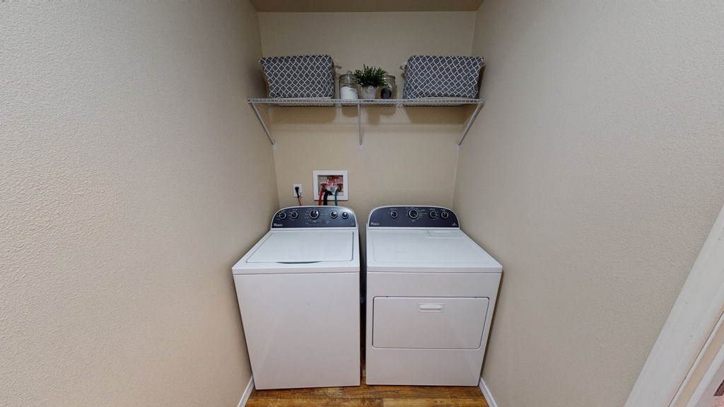 Laundry room here at Four Seasons Apartments with a washer and dryer side by side. Above them is a shelf with two patterned baskets and plants. The setting feels tidy and organized.