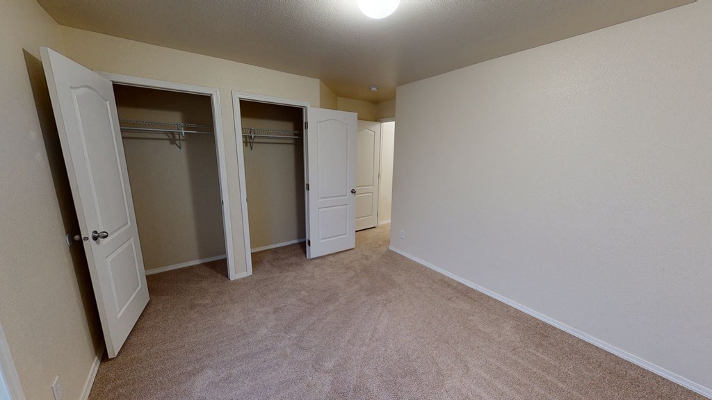 Bedroom here at Four Seasons Apartments with beige carpet and cream walls, featuring two open closets with white double doors, creating a spacious and neutral atmosphere.