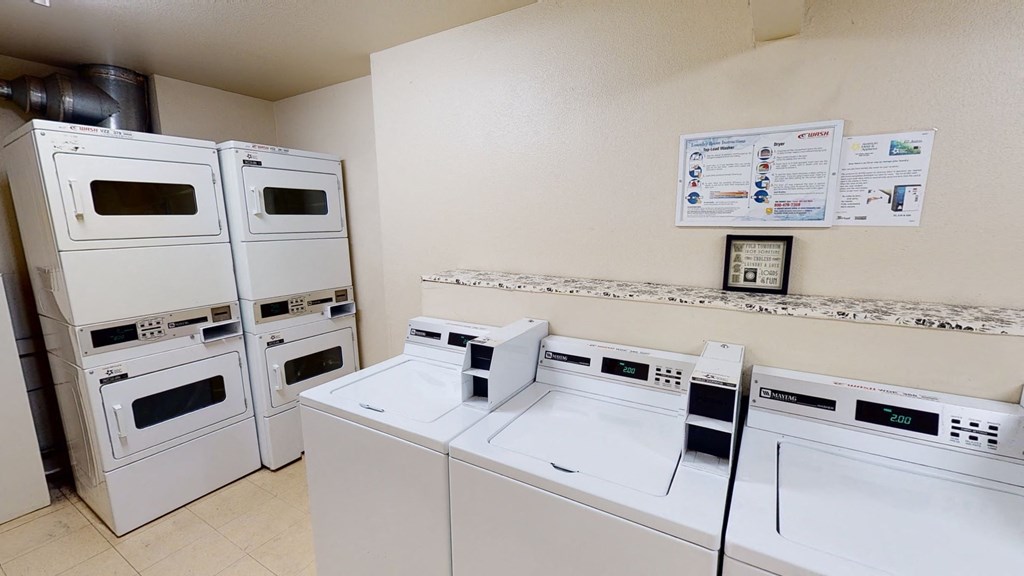 A clean laundry room here at Hillside Chalet Apartments with stacked dryers on the left and top-loader washers on the right. Instructions are mounted on a beige wall, creating an orderly atmosphere.