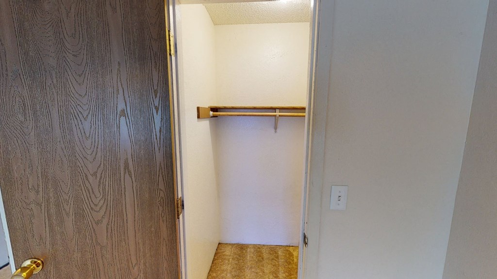 Open closet here at Rancho Tudor Apartments with wooden door ajar, revealing a white interior, a wooden hanging rod, and beige carpet. The space is minimal and tidy.
