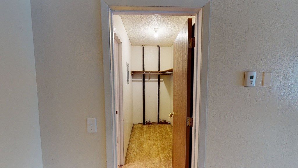 A small, empty closet here at Rancho Tudor Apartments with beige walls and carpet, an open wooden door, a single light bulb, metal rods, wooden shelves, and a wall thermostat.
