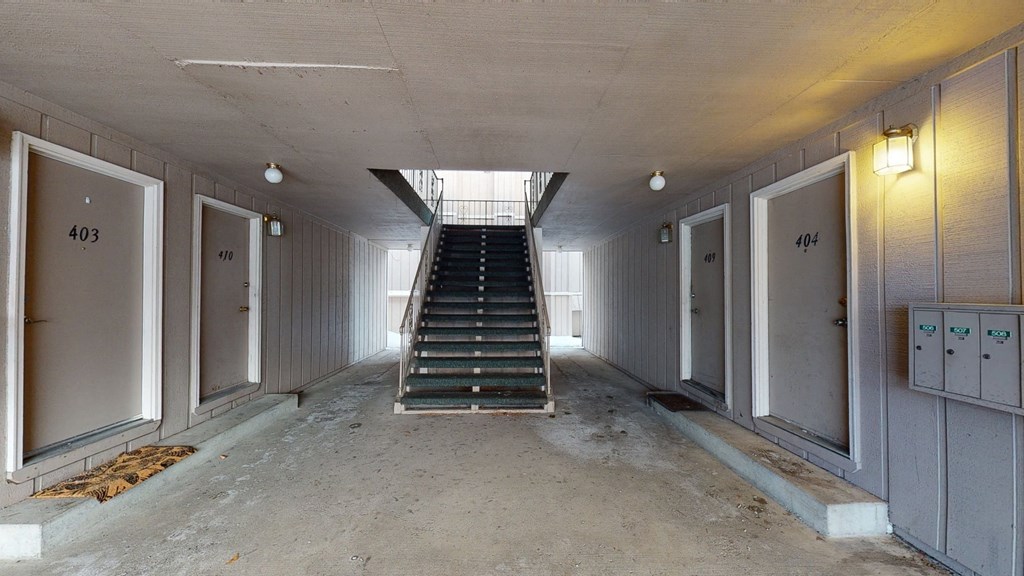 Apartment hallway here at Rancho Tudor Apartments with beige walls and closed brown doors numbered 403, 410, 409, 404. Central staircase leads to upper floor. Lit by small ceiling lights. Calm atmosphere.