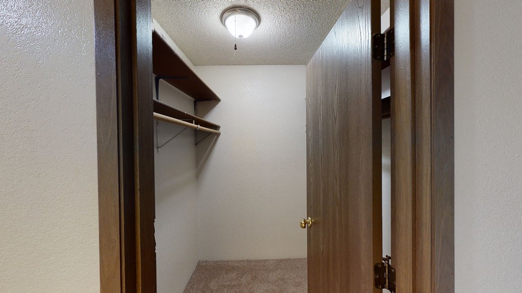 A small, empty walk-in closet here at Terrace on the Lake Apartments with beige carpet, white walls, and wooden shelves on the left. An open wooden door and a ceiling light create a cozy ambiance.