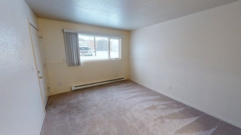 Empty living room here at The Club at Eagle Pointe Apartments with beige walls and carpet, featuring a large window with vertical blinds. Sunlight highlights the carpet's texture, creating a calm atmosphere.