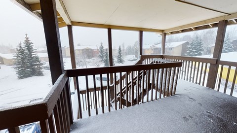 Snow-covered porch here at The Club at Eagle Pointe Apartments with wooden railings overlooks a snowy neighborhood. Fir trees and houses are visible under a gray, overcast sky. Quiet, wintry scene.