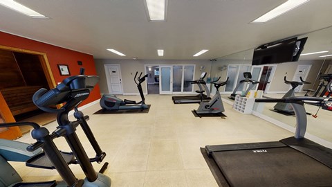 Bright gym room here at The Club at Eagle Pointe Apartments featuring treadmills, ellipticals, and stationary bikes on tiled floor. Mirrors and a flatscreen TV create a modern, spacious feel.