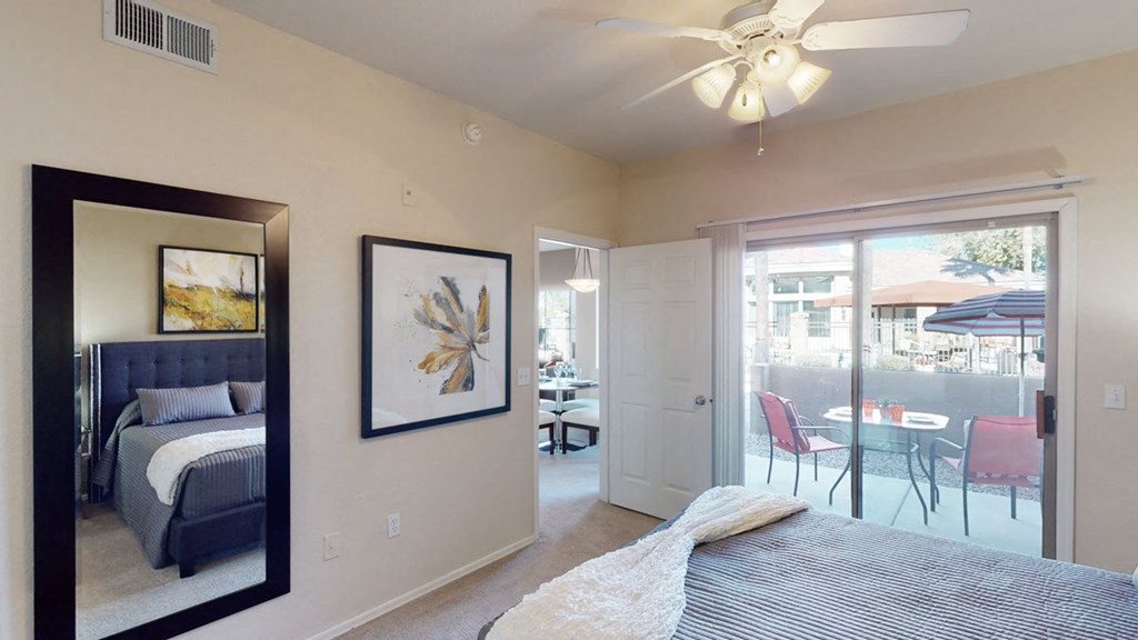 A cozy bedroom here at Dobson Towne Centre Apartments with a striped bedspread, mirror reflecting abstract wall art, and a ceiling fan. Sliding doors open to a patio with red chairs and an umbrella.