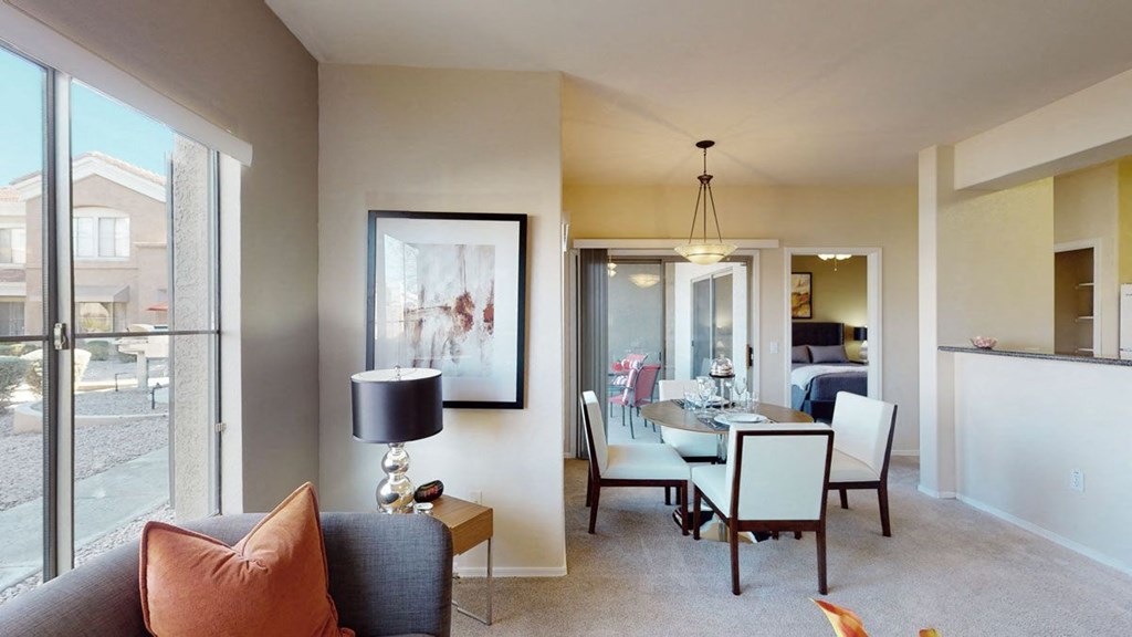 Modern living room here at Dobson Towne Centre Apartments with a grey couch and orange pillow, a framed abstract painting, and a lamp. Dining area visible with a round table, chairs, and soft lighting.