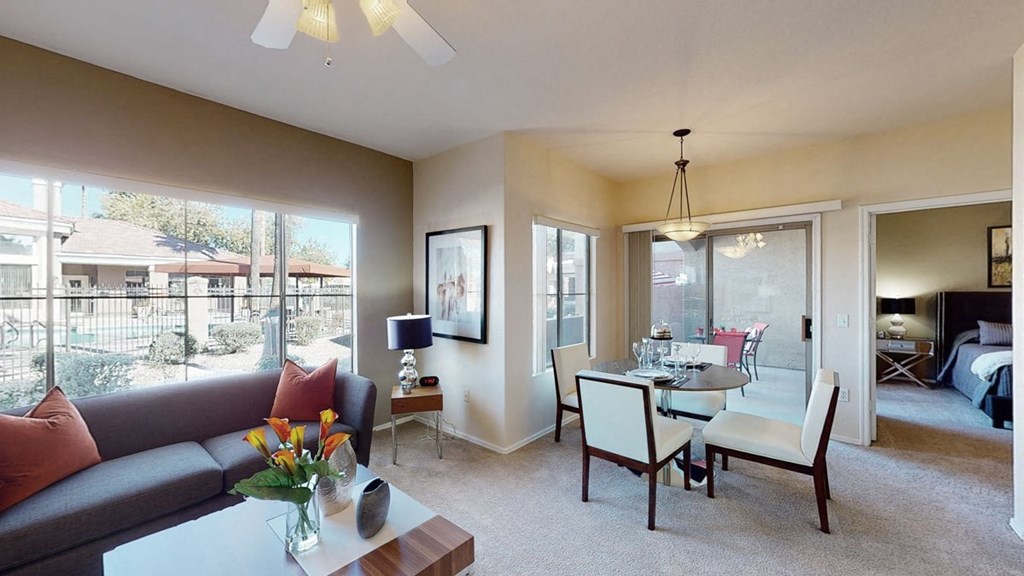 Modern living room here at Dobson Towne Centre Apartments with large windows overlooking a pool area. Gray sofa with orange pillows, glass-topped table with flowers, and dining area with white chairs. Bright and inviting atmosphere.
