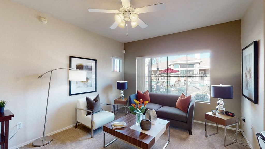 Contemporary living room here at Dobson Towne Centre Apartments with gray sofa, white armchair, and wood-accented furniture. Large windows overlook a patio. Neutral tones, cozy ambiance.