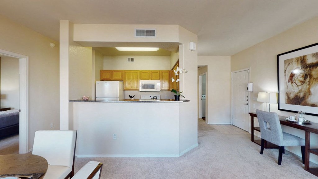 Bright, cozy apartment interior here at Dobson Towne Centre Apartments with an open kitchen featuring wooden cabinets, white appliances, and a small plant. Adjacent dining and living area with soft lighting creates a welcoming ambiance.