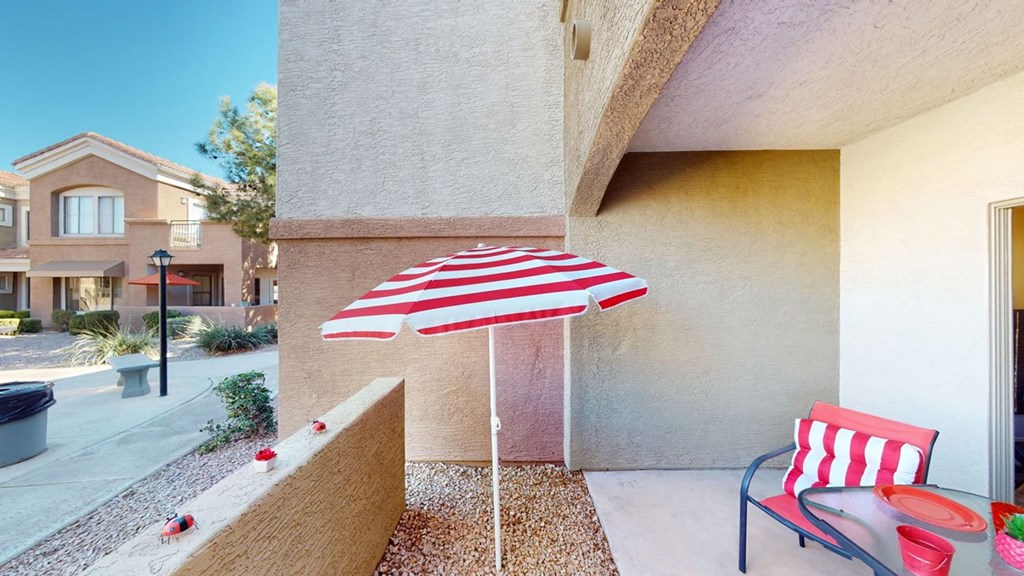 A sunlit patio here at Dobson Towne Centre Apartments features a red and white striped umbrella and matching chair cushion. Nearby, a glass table holds red items. A house and tree are visible in the background, conveying a cozy, cheerful vibe.