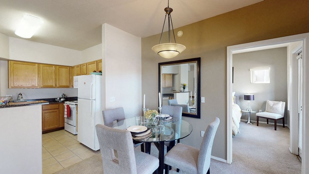 Dining area here at Dobson Towne Centre Apartments with glass table, four gray chairs, and a large wall mirror. Adjacent kitchen has light wood cabinets and white appliances. Bright, cozy atmosphere.