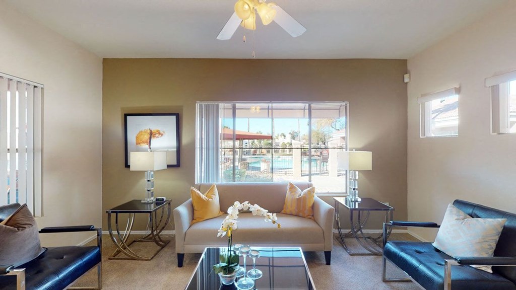 Stylish living room here at Dobson Towne Centre Apartments with a beige sofa, yellow pillows, and modern chairs facing a glass coffee table. Large window reveals a sunny poolside view. Neutral tones.